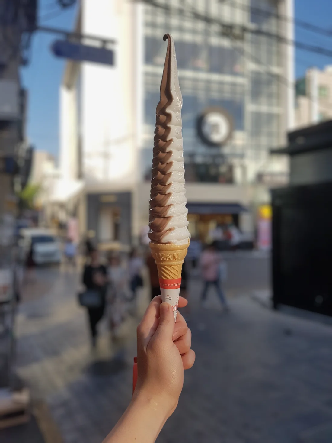 A hand holding a tall chocolate and vanilla 32Parfait soft serve on a street in Hongdae, Seoul